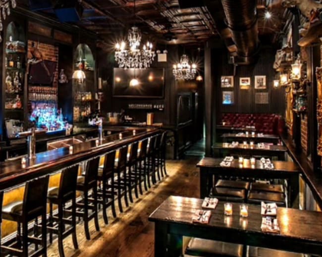 The image shows the interior of a bar with a long wooden counter, high stools, and several tables arranged in a dimly lit atmosphere adorned with chandeliers and dark wood paneling.