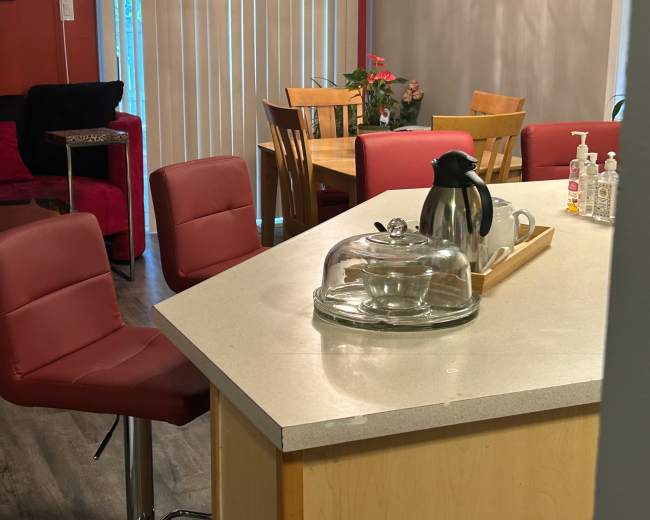 The image shows a kitchen area featuring a countertop with a glass serving dome, a coffee pot, and several red chairs arranged around the space.