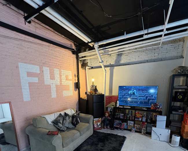 The image shows a cozy basement living area featuring a light-colored sofa, a large TV mounted on the wall, and a variety of furnishings and decorations against a textured pink wall.
