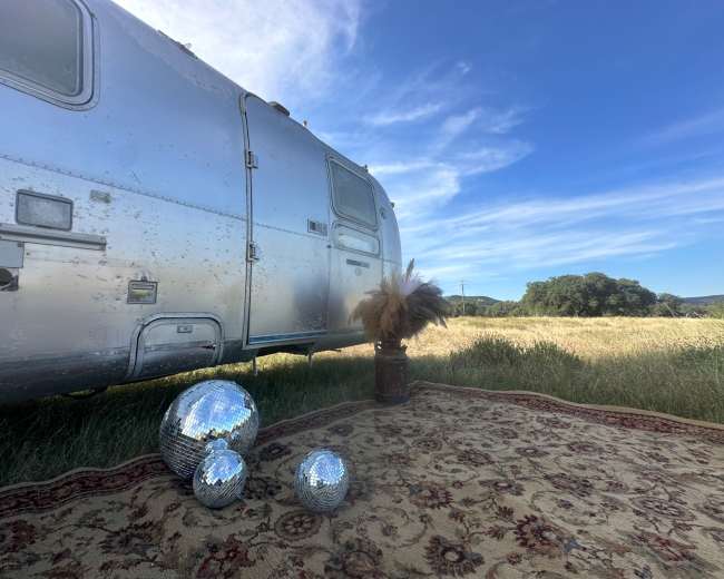 An Airstream trailer is parked beside a rug, with three disco balls and a decorative plant nearby, set against a backdrop of a grassy field and blue sky.