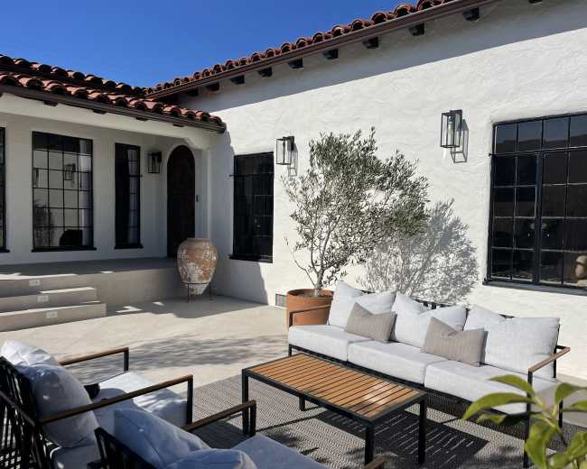 The image shows a patio area featuring modern outdoor furniture, a decorative pot, and a small tree beside a white stucco building with black window frames.