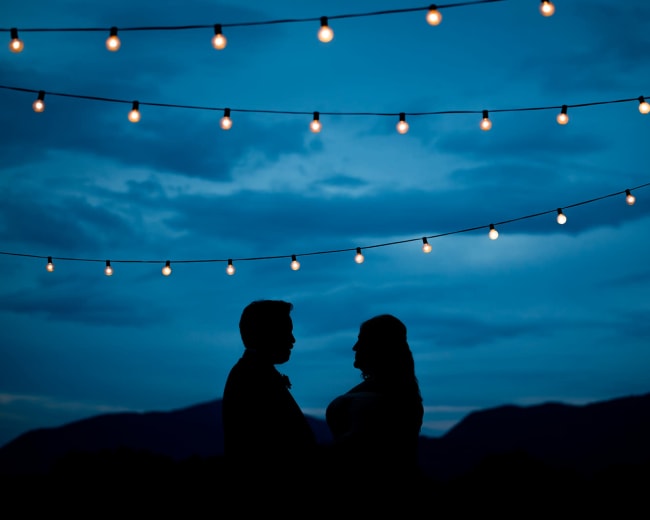 A couple stands in silhouette under string lights against a twilight sky with mountains in the background.