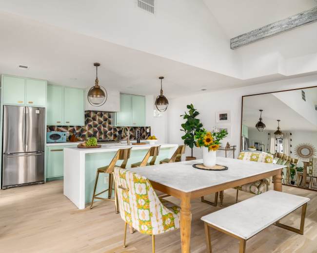 The image shows a modern kitchen and dining area featuring mint green cabinets, a large table with floral-patterned chairs, and bright sunlight streaming through a window.