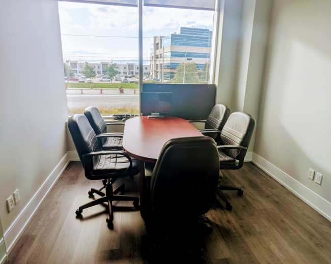The image shows a small conference room with a round table and four black chairs near a large window, overlooking a street and buildings outside.