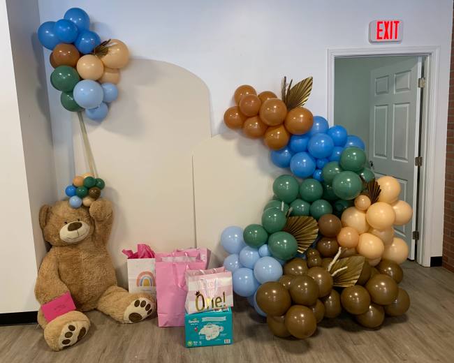 A large teddy bear seated next to a cluster of colorful balloons and gift bags arranged for a celebration.