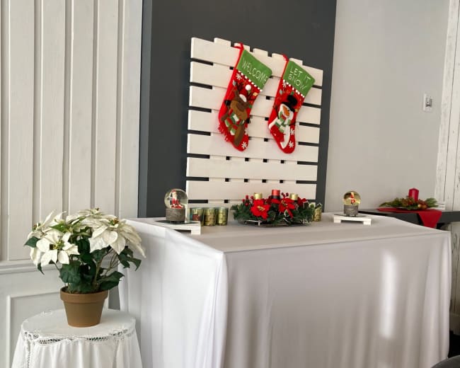 A decorated table with a white cloth displays holiday stockings and floral arrangements against a gray wall.