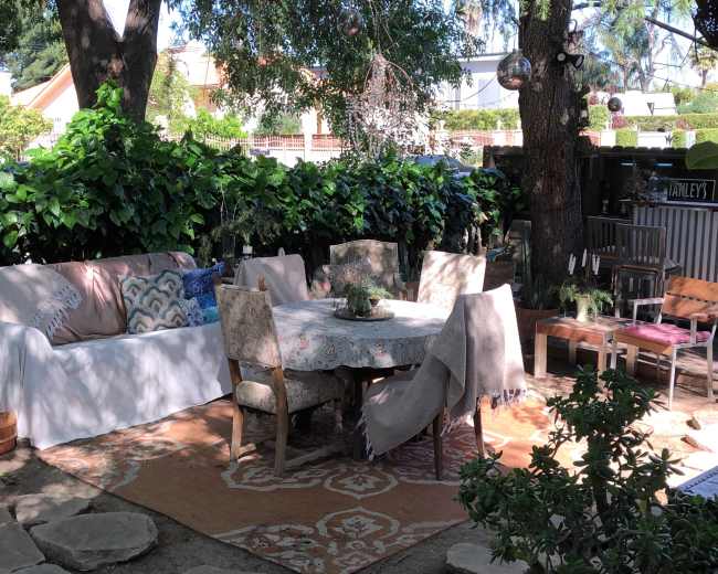 The image shows an outdoor seating area with a round table surrounded by chairs, a large white sofa, and greenery in the background.