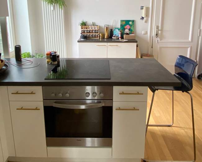 A modern kitchen features a central island with a stovetop, a chair, and various kitchen appliances and decor against a neutral background.