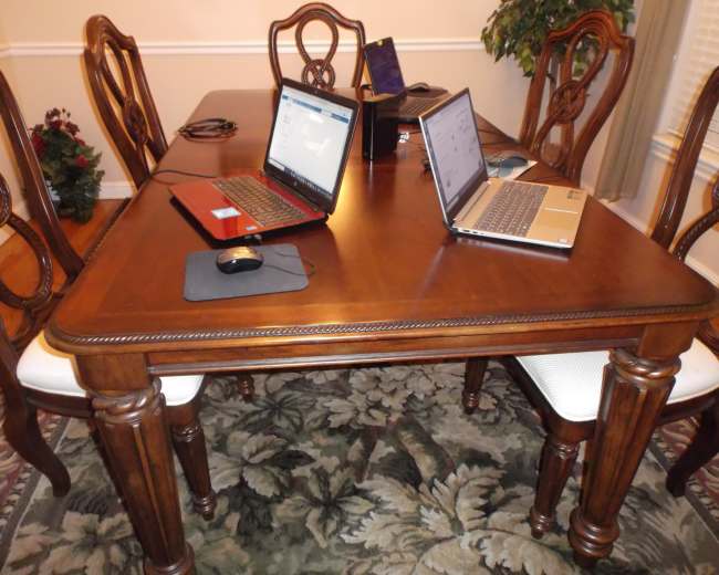 A wooden dining table is set with two laptops and a mouse, surrounded by chairs on a patterned rug.