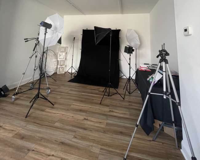 A photography studio setup with a black backdrop, lighting equipment, and tripods arranged in a spacious room with wooden flooring.