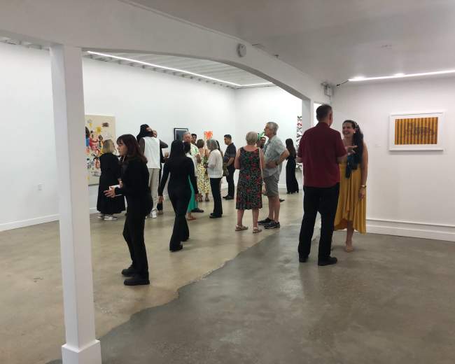 A group of people is gathered in a spacious art gallery, engaging in conversation while observing various artworks on the walls.