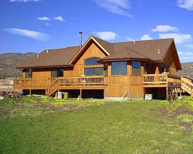 A large wooden house with multiple gabled roofs and a spacious deck overlooks a grassy yard against a mountainous backdrop.