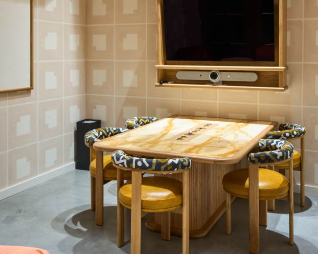A small conference room features a wooden table with four chairs, a wall-mounted television, and patterned wallpaper.