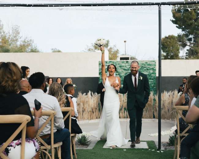 A bride and groom walk hand in hand down the aisle, with the bride holding a bouquet and raising her arm in celebration.