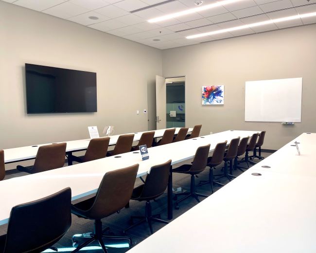 A conference room features long tables with brown chairs, a wall-mounted screen, a whiteboard, and an abstract artwork.