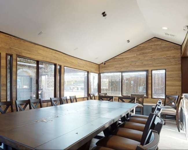 The image shows a spacious conference room with a long wooden table surrounded by chairs, large windows featuring greenery outside, and wood-paneled walls.