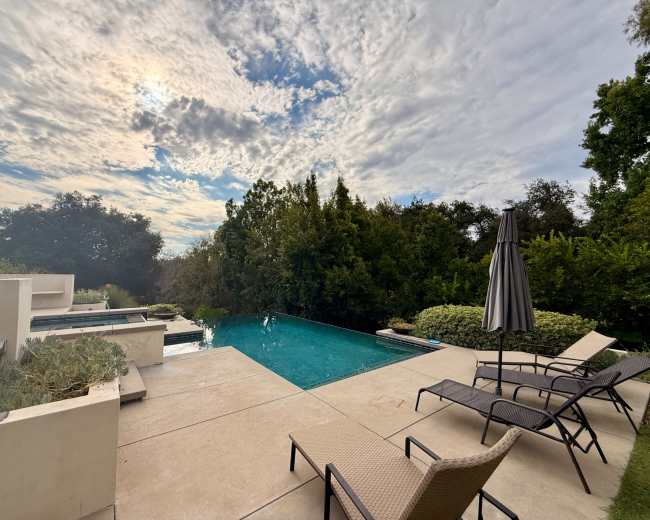 The image shows a tranquil backyard scene featuring a swimming pool, lounge chairs, and lush greenery under a cloudy sky.