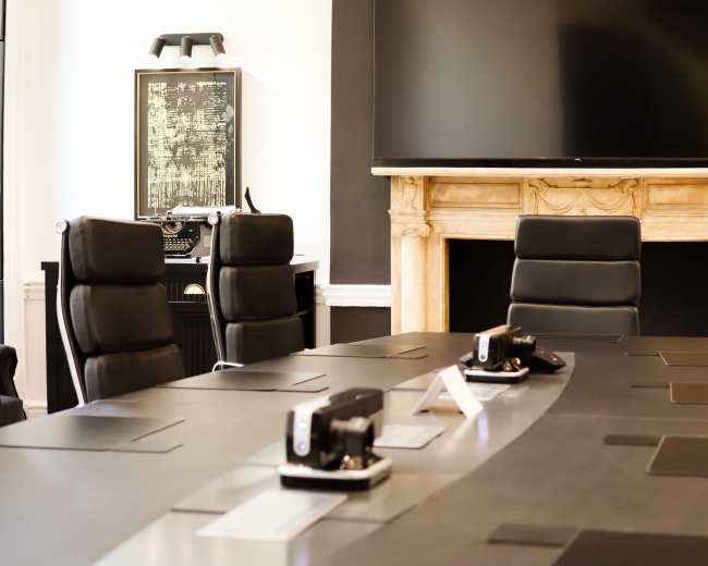 The image shows a modern conference room with a long table, black leather chairs, and a large screen mounted on the wall.