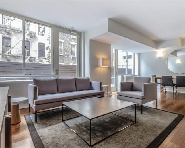 The image shows a modern living room featuring two gray sofas, a glass coffee table, and a dining area with a wooden table and black chairs, all in a bright space with large windows.