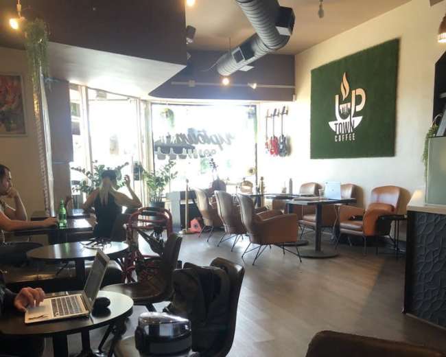 The image shows a modern coffee shop with a mix of seating options, including tables and armchairs, and a clear view of customers working on laptops.