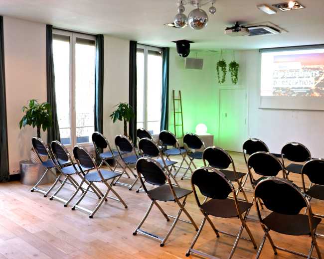A seminar room is set up with rows of black folding chairs facing a screen, illuminated by a green light, with plants and a ladder in the background.