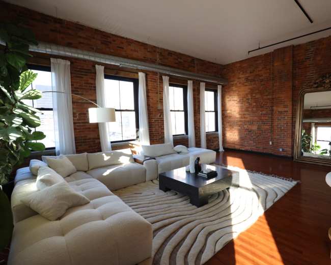 The image shows a spacious living room with exposed brick walls, large windows, and a modern sectional sofa centered around a dark coffee table on a light rug.