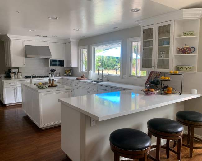 A modern kitchen features white cabinets, a large island with bar stools, and windows that provide natural light and a view of the outdoors.