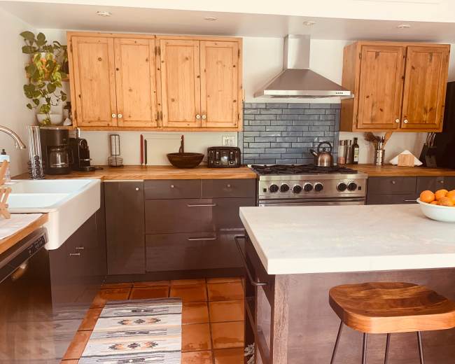 The image shows a modern kitchen with wooden cabinetry, stainless steel appliances, and a central island topped with a bowl of oranges.