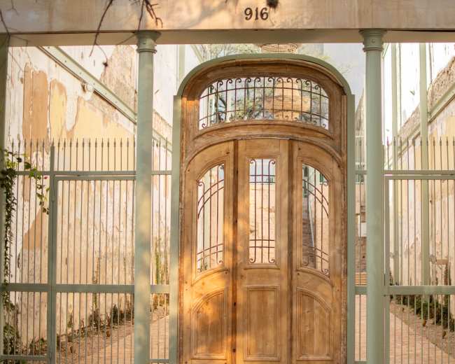 The image shows an intricately designed wooden gate with wrought iron accents, set within a weathered stone enclosure and flanked by pillars.