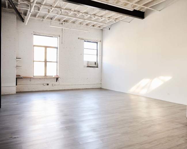 A spacious, empty room with white walls and wooden flooring features a window allowing natural light and an air conditioning unit mounted nearby.