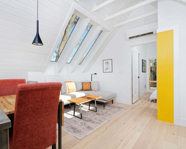 The image shows a bright, modern living space with sloped ceiling, large skylights, a dining area with red chairs, a gray sofa, and light wood flooring.