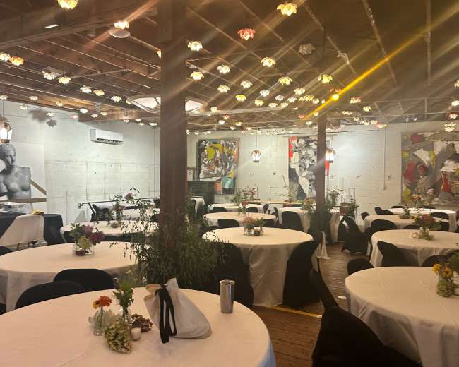 The room features several round tables covered with white tablecloths, surrounded by black chairs, and adorned with floral centerpieces, while artwork and decorative lights enhance the space.