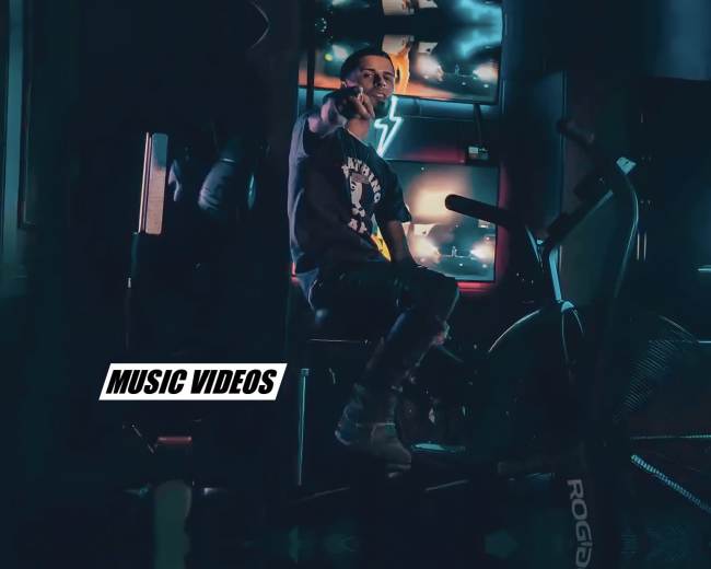 A person sits on an exercise bike in a dimly lit room with neon lights and a television displaying music videos in the background.