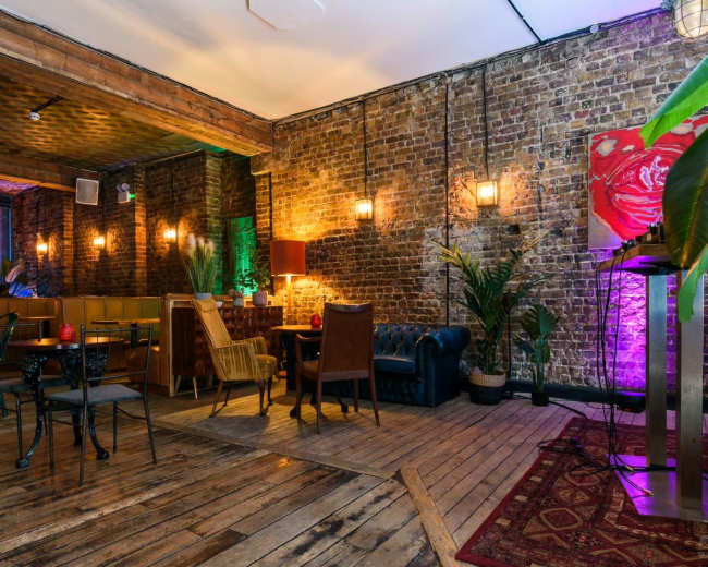 The image displays a rustic indoor space featuring exposed brick walls, wooden floors, and a variety of seating arrangements, including a blue leather sofa and tables with chairs, accented by plants and soft lighting.