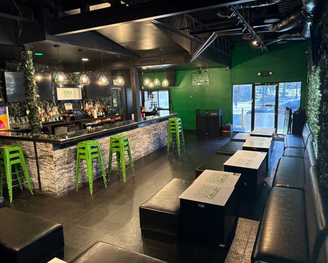 The image shows a modern bar area with a black and green color scheme, featuring a sleek bar counter, high stools, and seating areas with low tables.