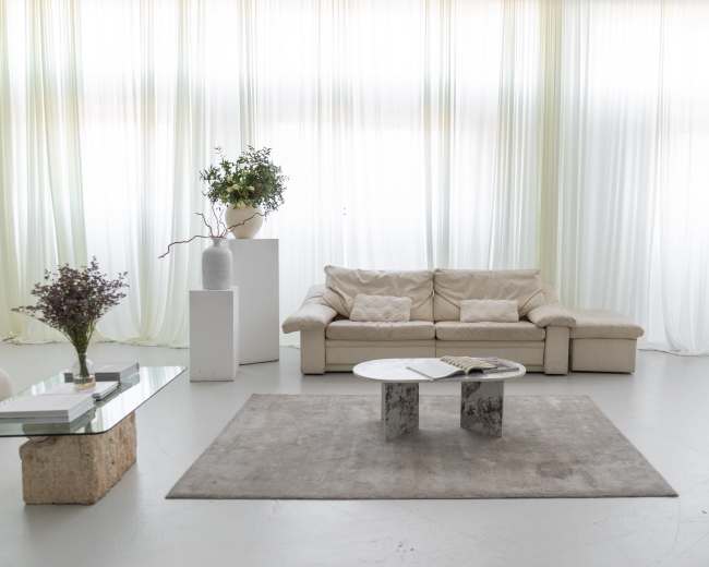 A modern living room features a beige sofa, a glass coffee table, and a light gray rug under large sheer curtains.