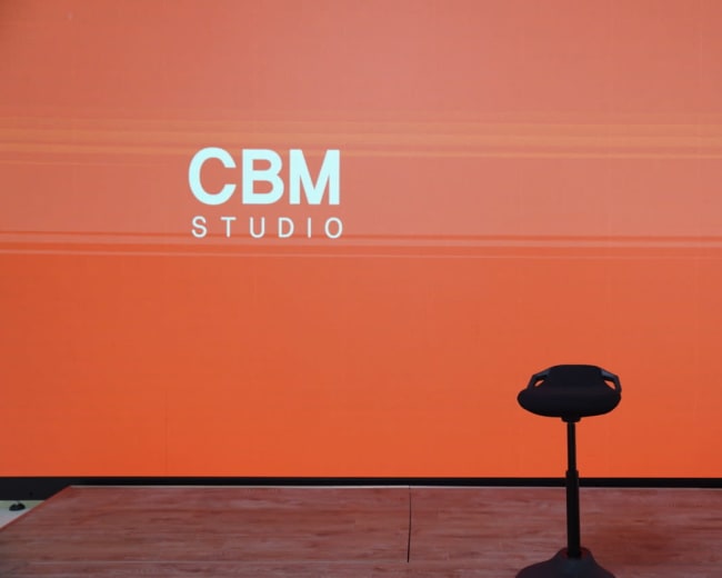 The image shows an empty stage area with a simple black stool in front of a large orange backdrop displaying the text "CBM STUDIO."