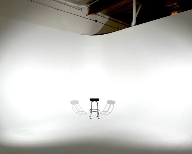 A black stool casts a shadow on a plain white backdrop in an empty studio space.