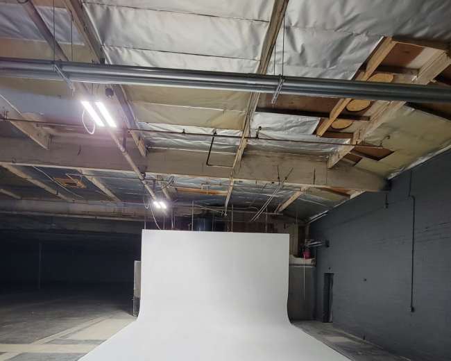 A large, white seamless backdrop is set up in a dimly lit industrial space with exposed beams and concrete flooring.