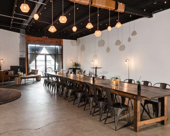 The image shows a spacious dining area with a long wooden table, metal chairs, and warm hanging lights illuminating the neutral-toned space.