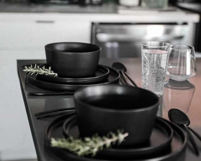 A dark kitchen countertop is set with two black bowls, black plates, and utensils, garnished with sprigs of rosemary, alongside a glass of water and a clear tumbler.
