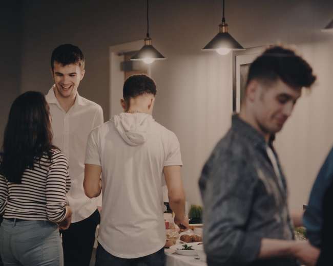 A group of people engages in conversation at a social gathering near a buffet table in a well-lit room.