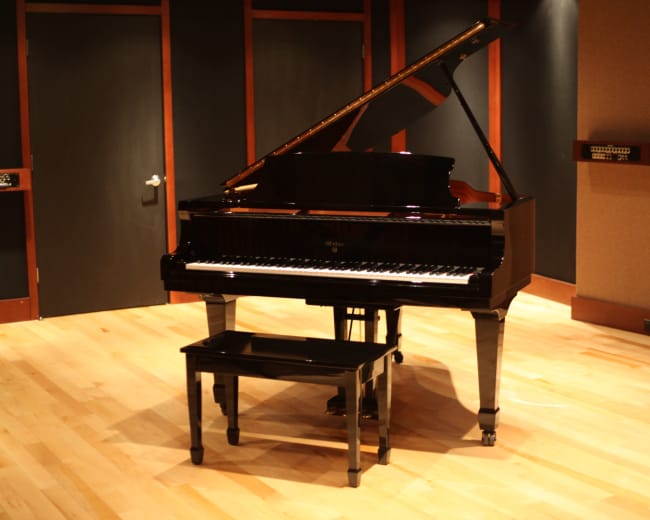 A grand piano is positioned on a wooden floor in a soundproof room with dark walls and a music stand.