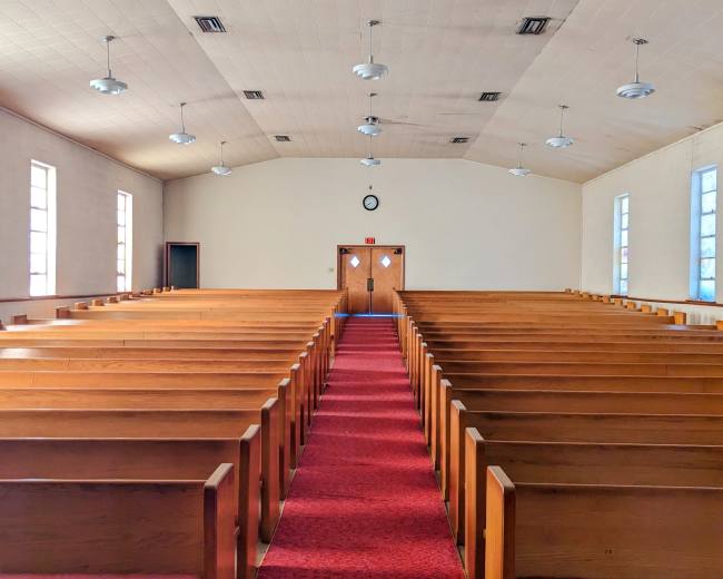 The image shows the interior of a church with rows of wooden pews facing a central aisle that leads to a set of double doors at the rear.