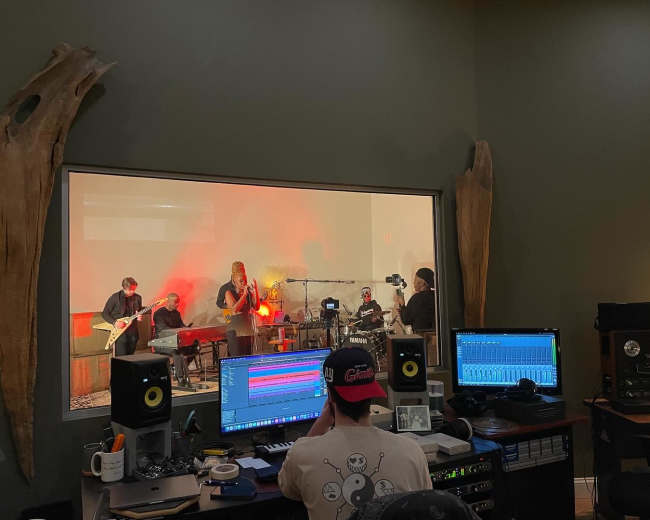 A band performs in a studio behind a glass wall, while a person works on a music production setup in the foreground.