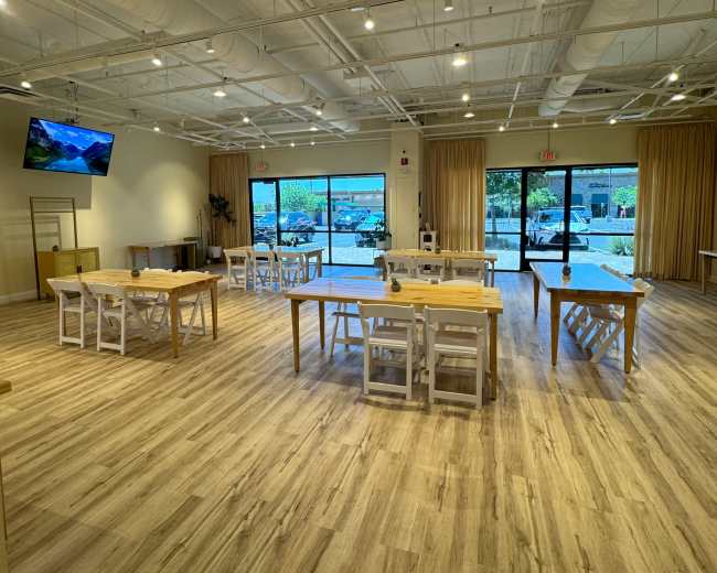 A spacious, well-lit interior features several wooden tables and chairs arranged neatly on a wooden floor, with large windows showcasing an outdoor view.