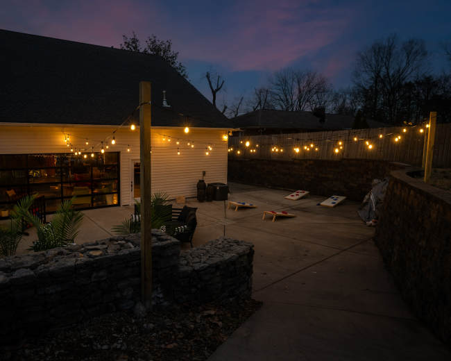 The image shows a backyard patio with string lights illuminating a concrete area, flanked by stone walls and a garage in the background.