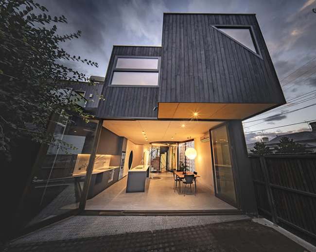 A modern, angular house with a dark wood exterior is shown against a twilight sky, featuring large windows and a spacious open interior connected to an outdoor patio.