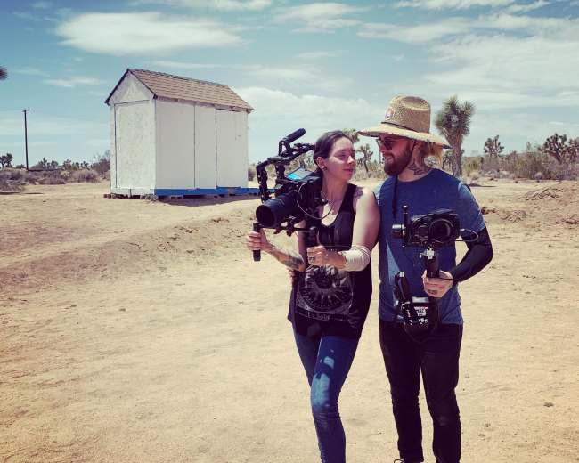 Two individuals hold video cameras while standing in a desert landscape near a small white shed.
