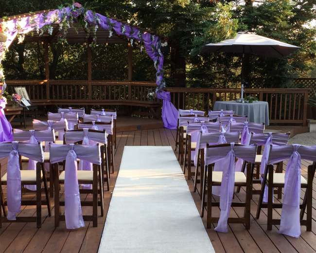 A wedding ceremony setup on a wooden deck, featuring rows of chairs adorned with purple sashes and a white aisle runner leading to a floral arch.